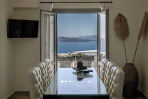 A dining area with a glass table, white upholstered chairs, a television on the wall, and a view of the sea.