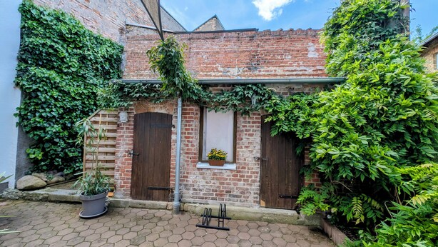 Un bâtiment en briques avec deux portes en bois, une fenêtre agrémentée d’une jardinière et un pot de fleurs posé sur le sol.