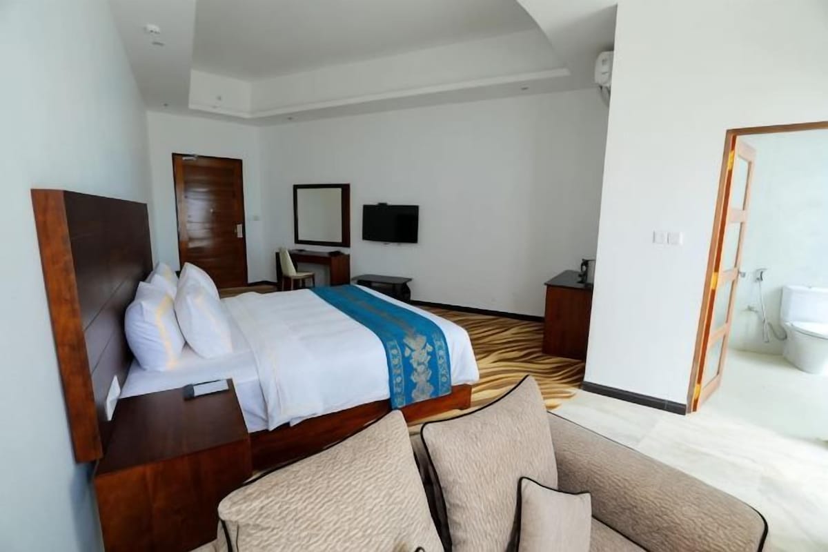 Une chambre d’hôtel avec un lit, un canapé, une télévision, un bureau et une salle de bain visible à travers une porte ouverte.