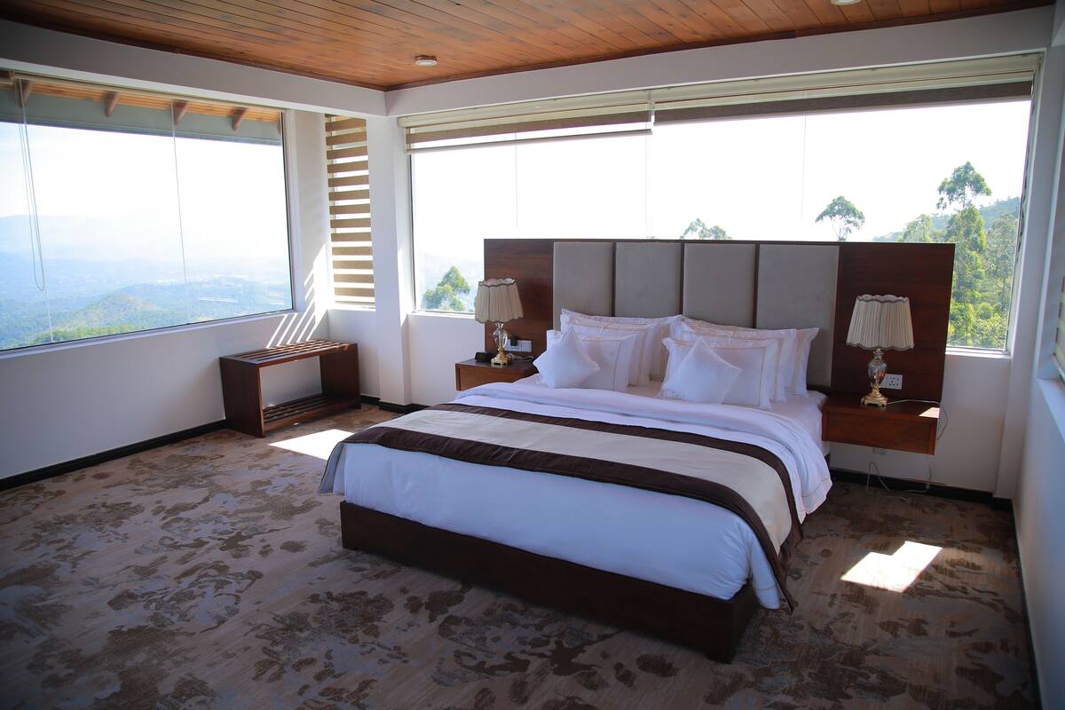 A large bed with a wooden headboard, two bedside tables with lamps, and a view of the mountains through large windows.