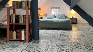 Une chambre mansardée chaleureuse avec un plafond en pente, une bibliothèque remplie de livres, un lit avec des oreillers et des carreaux de sol à motifs.