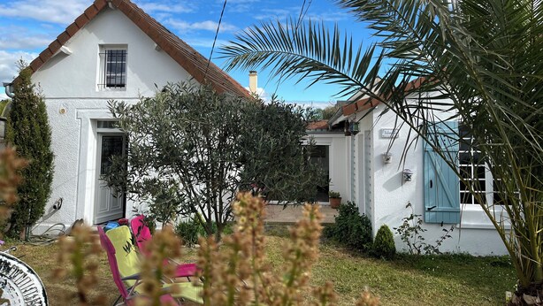 Une maison blanche avec un toit marron, une petite fenêtre et une porte. Il y a un jardin avec différentes plantes et une chaise.