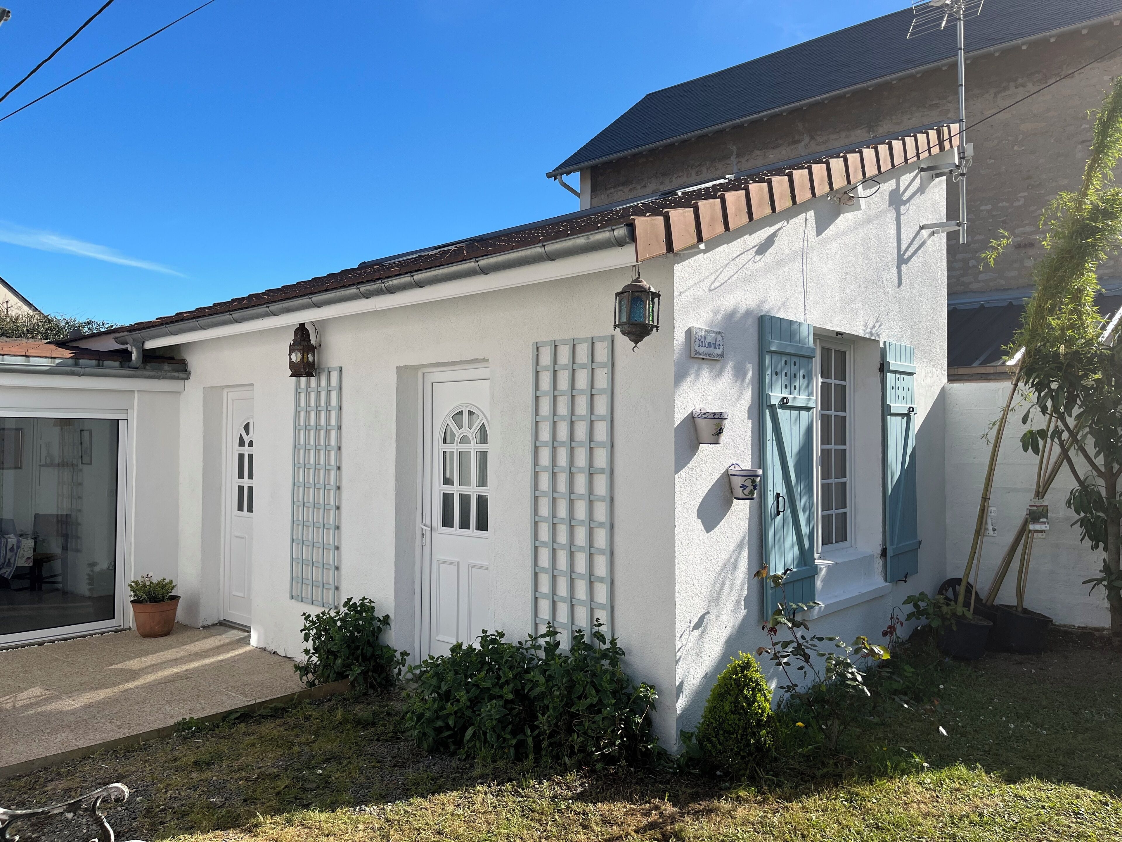 A white building with a tiled roof, a front door, and a window with shutters. There is a potted plant and a small garden area in front of the building.