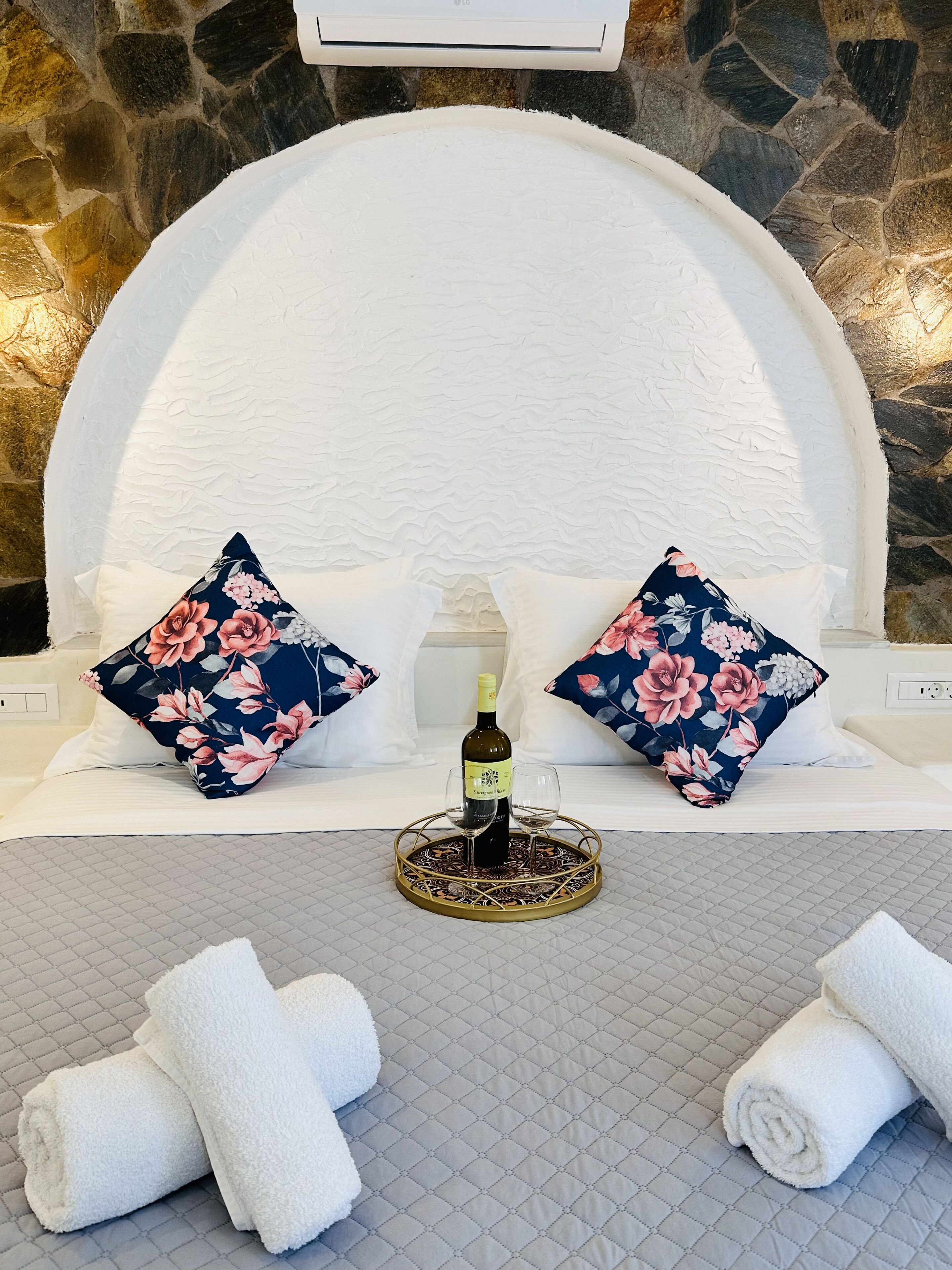A bed with floral pillows, a wine bottle, and glasses on a tray, with rolled towels on the floor.