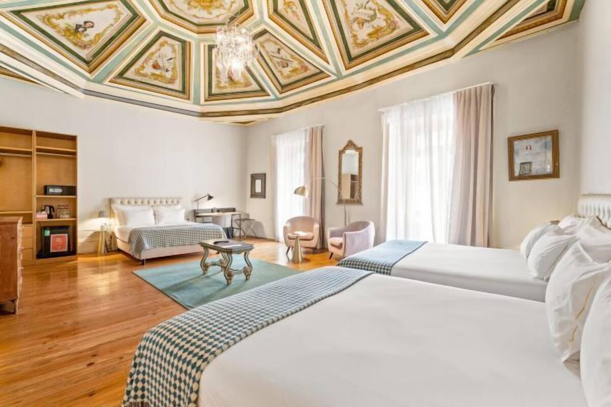 A hotel room with two beds, a chandelier, a wooden floor, and a ceiling with a geometric pattern.