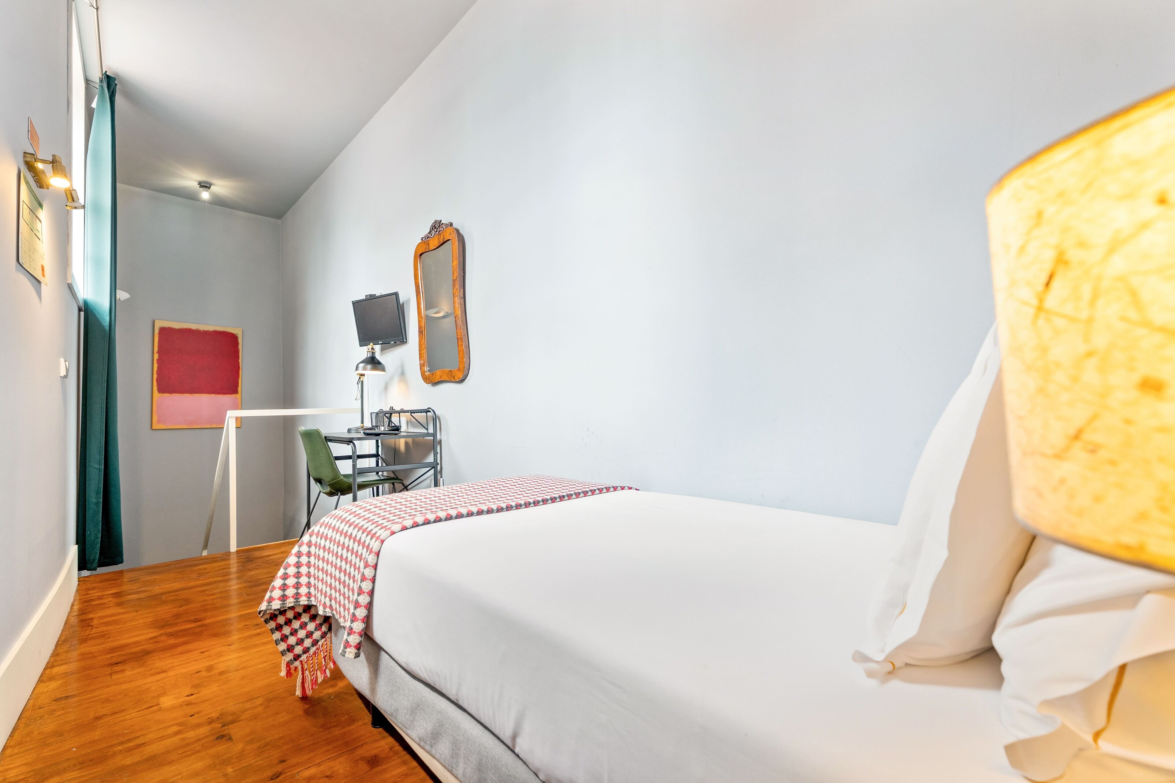 A bedroom with a bed, a desk, a chair, a mirror, and a red artwork on the wall.