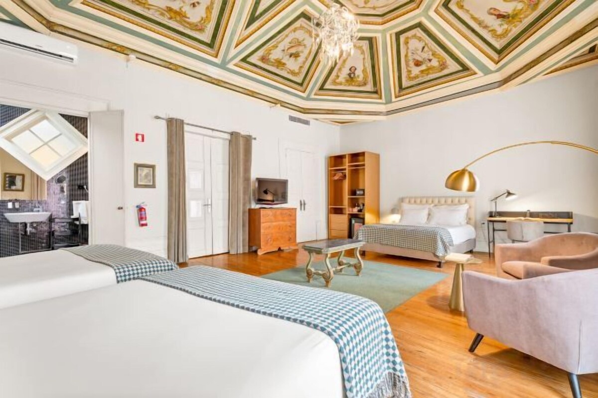 A hotel room with a large bed, a smaller bed, a desk, a chair, a TV, and a ceiling with intricate designs.