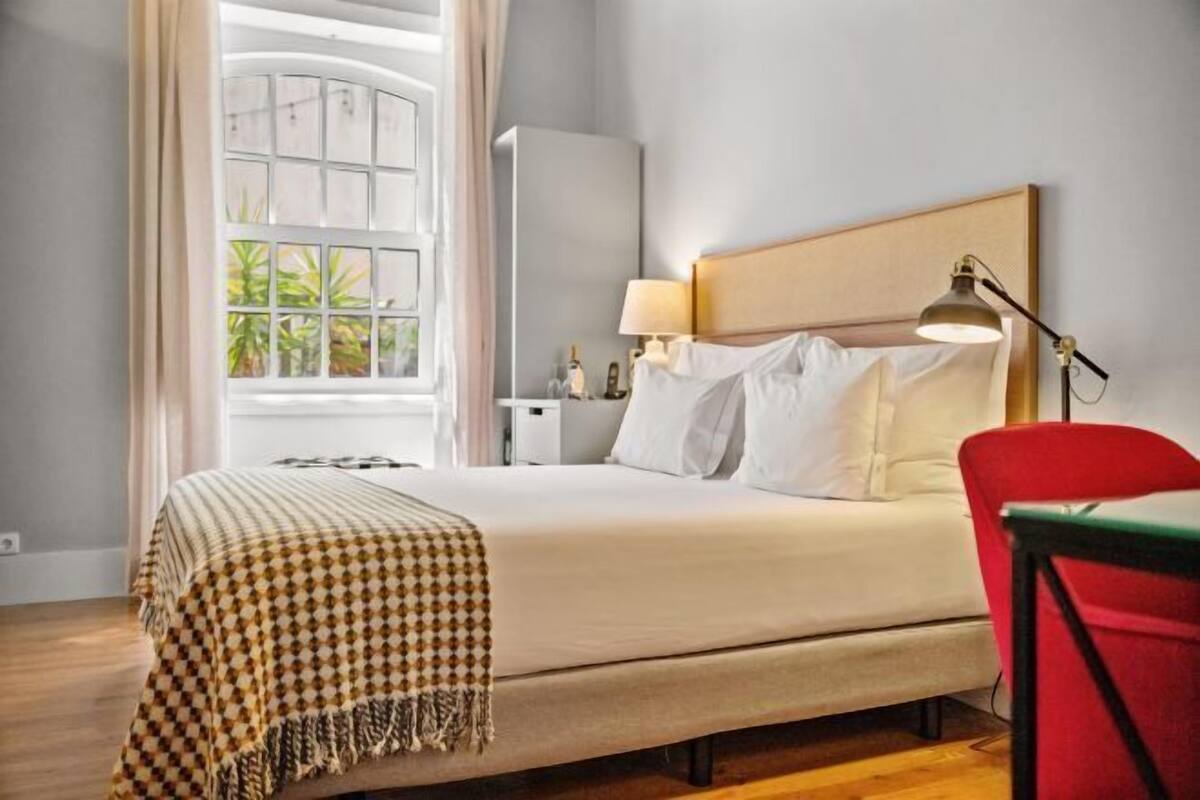 A hotel room with a large bed, a bedside table with a lamp, a window with curtains, and a red chair.