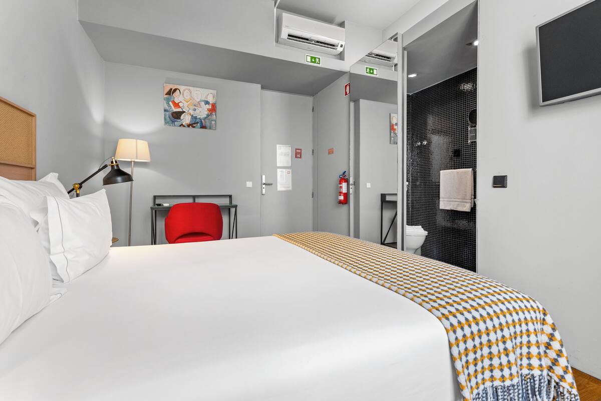 A hotel room with a bed, a red chair, a lamp, an air conditioning unit, and a shower area.