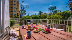 A wooden deck with a table set for breakfast, surrounded by a white fence, with a view of a residential building and greenery in the background. - Apartment near the beach Avus in Alcudia (Alcúdia)