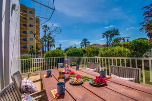 A wooden deck with a table set for breakfast, surrounded by a white fence, with a view of a residential building and greenery in the background.