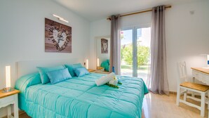 A bedroom with a large bed, a desk, and a chair. There are two windows with curtains, and a view of greenery outside. - Apartment near the beach Avus in Alcudia (Alcúdia)