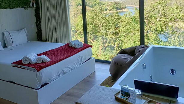 A modern bedroom with a large bed, a view of the outdoors, a massage chair, and a small table with amenities. - Dobau Village (Vieira do Minho)