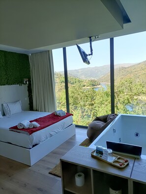 A modern bedroom with a large bed, a view of the outdoors, a massage chair, and a small table with amenities. - Dobau Village (Vieira do Minho)