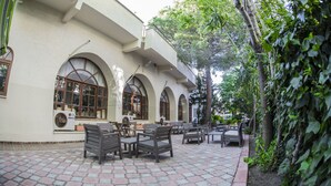 Un espace extérieur aménagé avec des tables et des chaises, un bâtiment doté de fenêtres en arc de cercle et un jardin avec des arbres et des plantes.