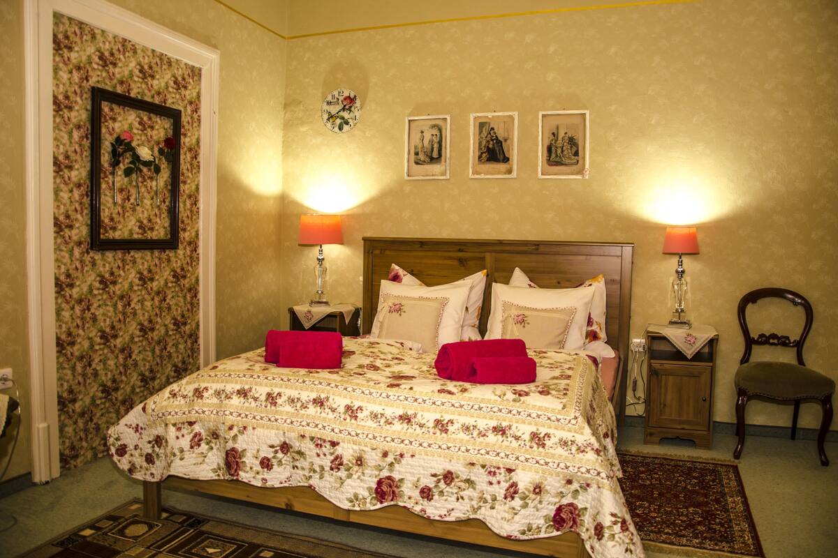 Une chambre à coucher avec une couvre-lit à motifs floraux, deux lampes de chevet, une tête de lit en bois et une petite table de chevet en bois avec une lampe.