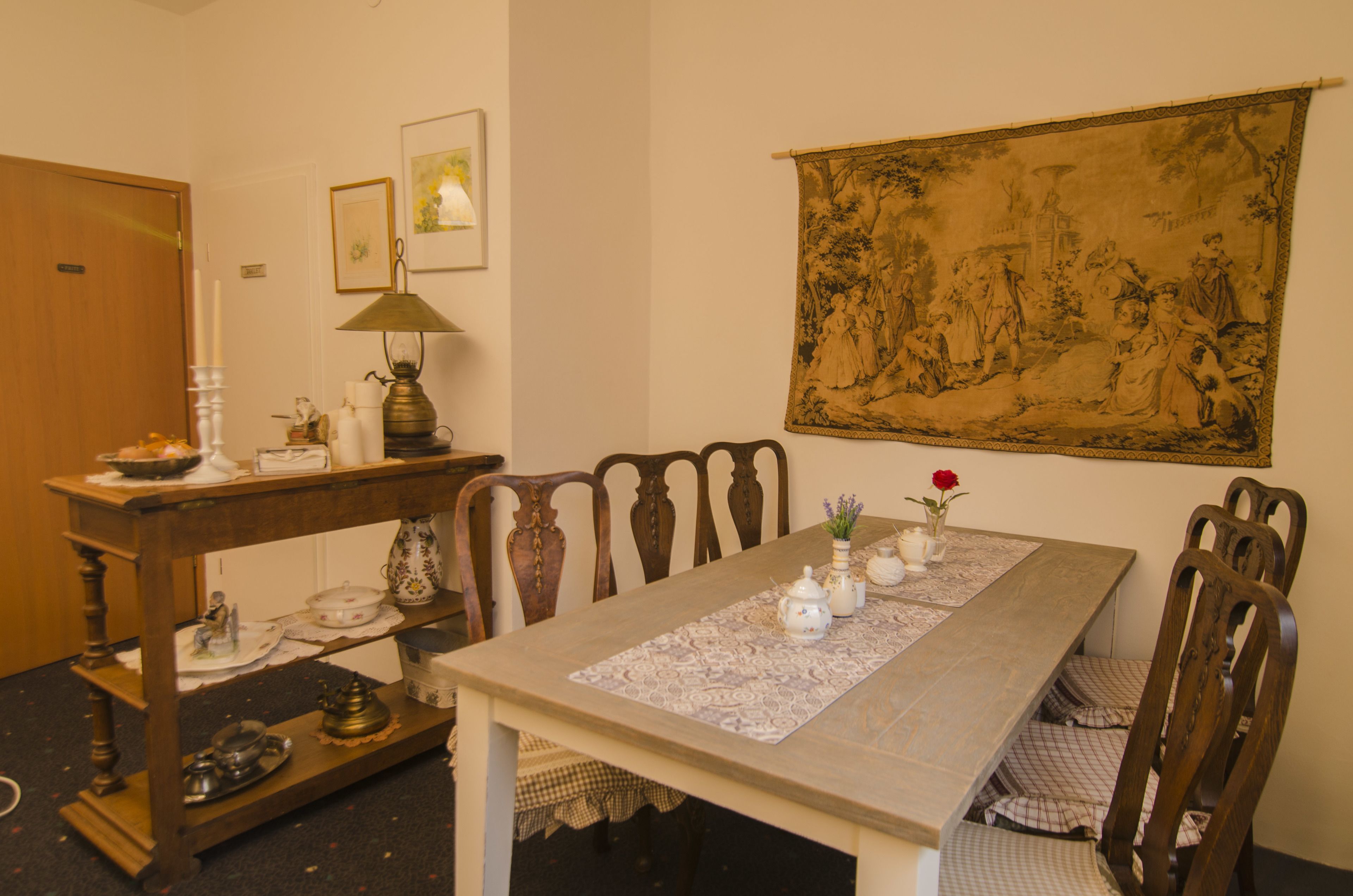 A dining area with a wooden table, chairs, a lace table runner, a tapestry on the wall, and a lamp.