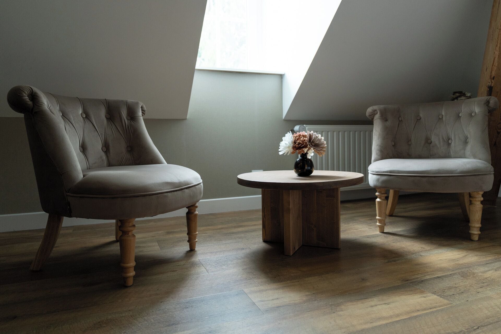 Una habitación con dos sillones tapizados, una pequeña mesa de madera con un jarrón de flores y un techo inclinado con tragaluz.
