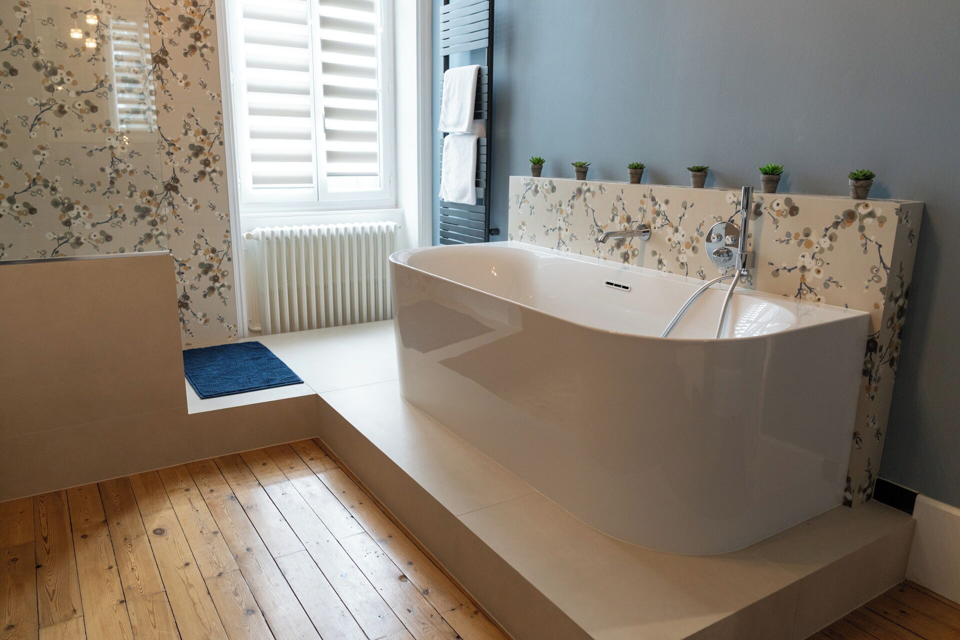 A modern bathroom with a freestanding bathtub, a radiator, and a window with shutters.