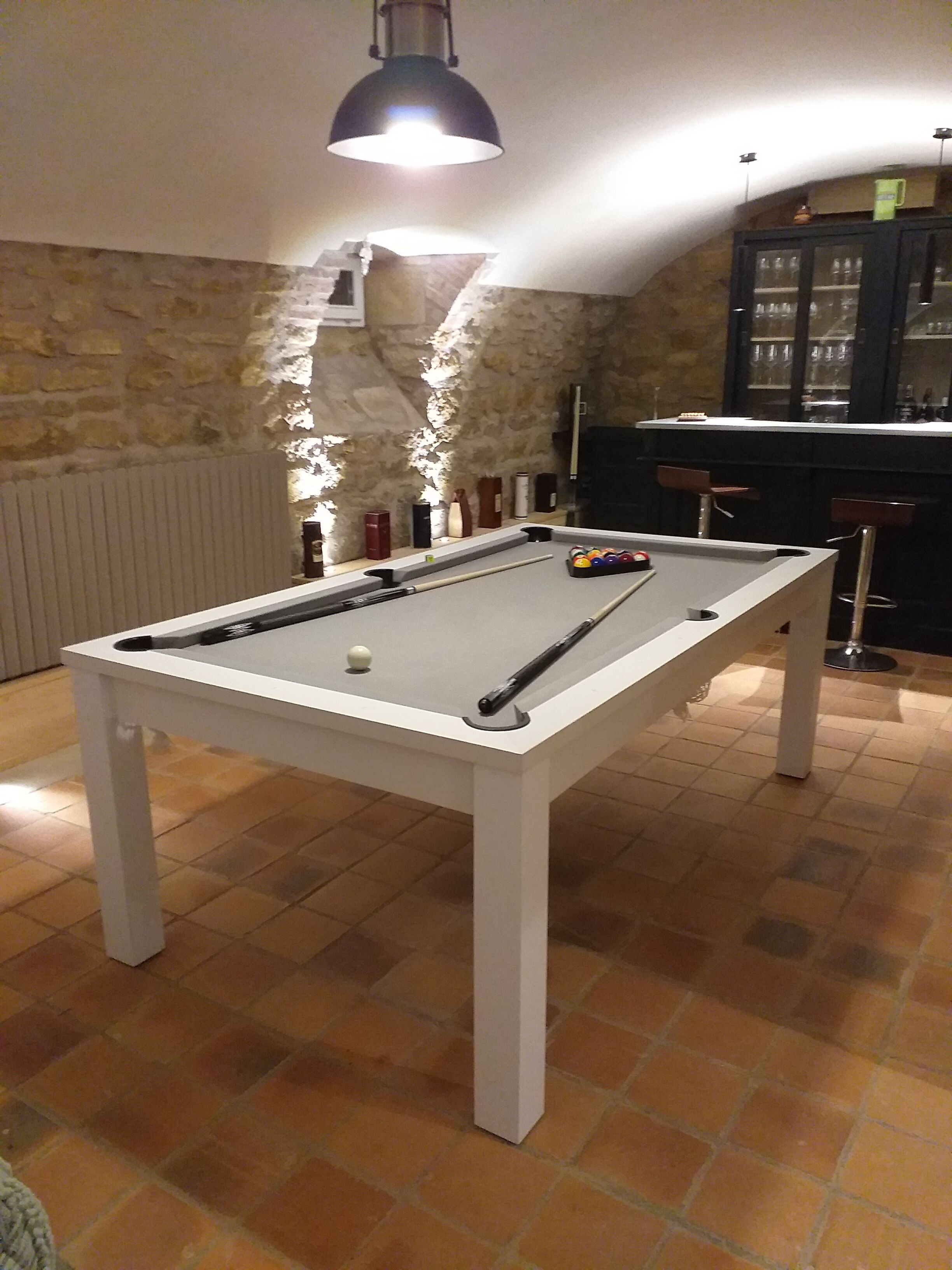 Uma mesa de bilhar com bolas e tacos, uma área de bar com banquetas e uma parede de pedra.
