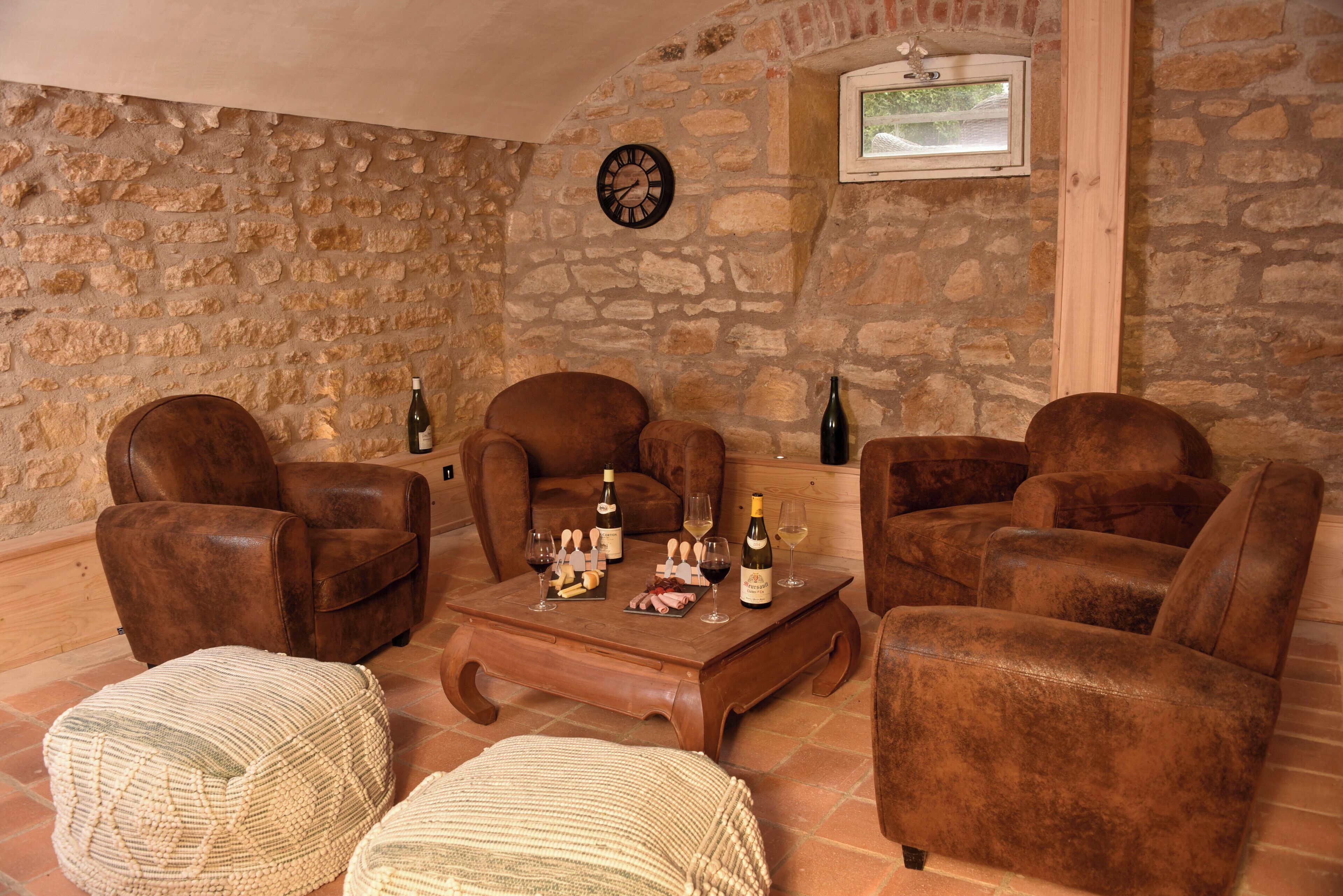 A cozy room with stone walls, leather armchairs, a wooden coffee table with wine bottles and snacks, and a clock on the wall.