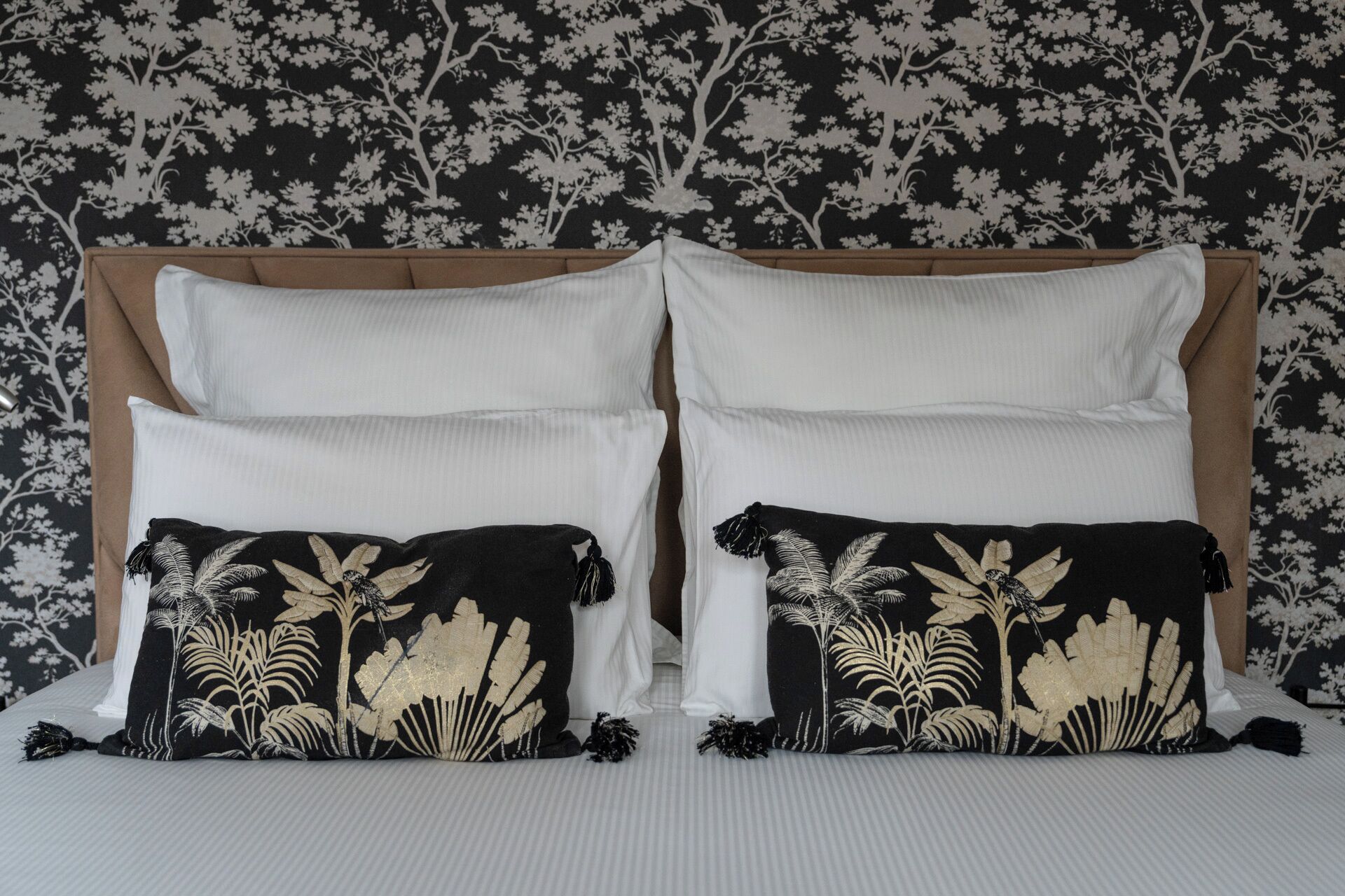 Un letto con lenzuola bianche, due cuscini neri con decorazioni in foglia d'oro e una testiera con motivo floreale.