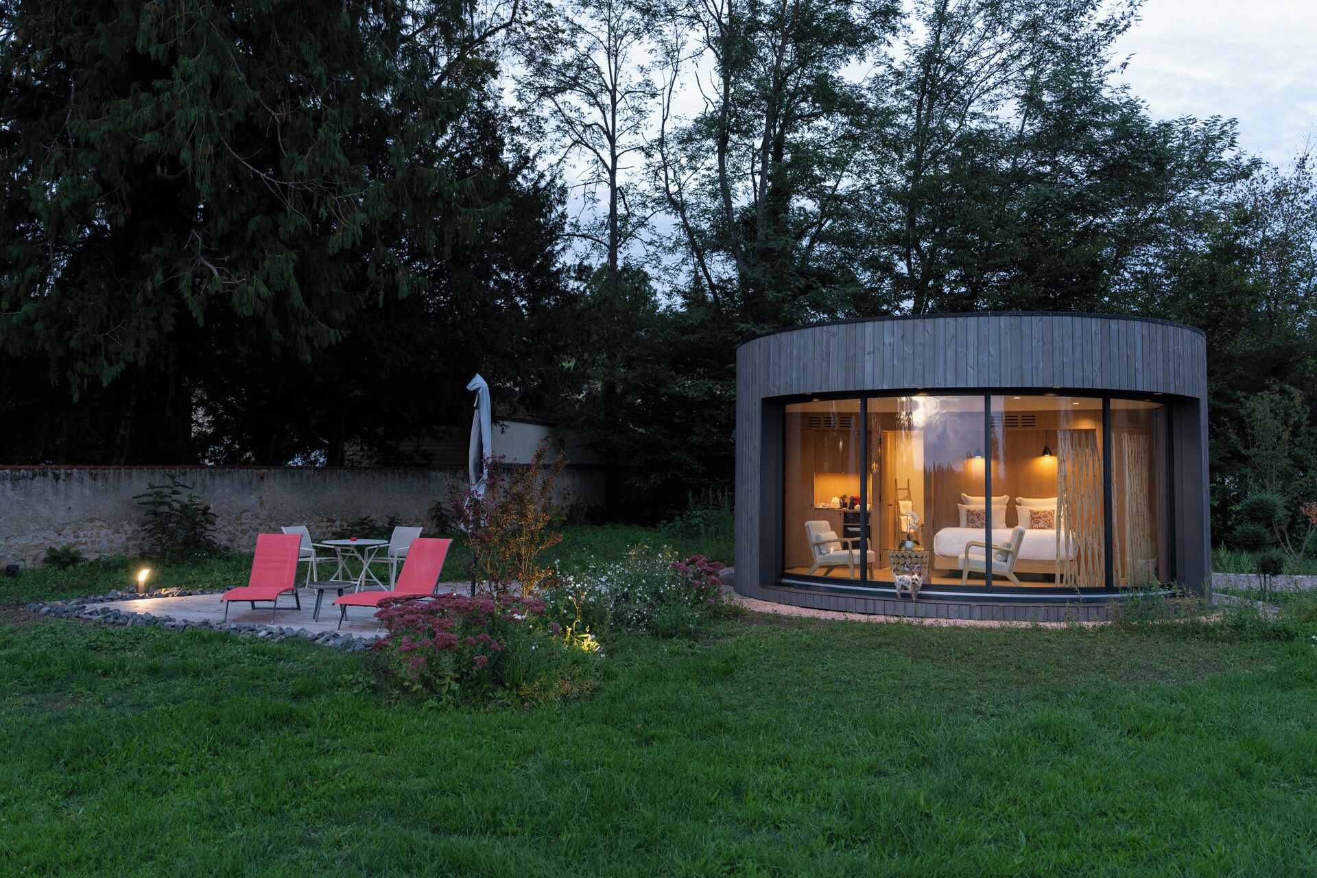 A modern, circular glass structure with a bed and seating area, surrounded by a garden and trees.