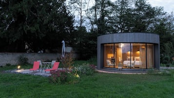 A modern, circular glass structure with a bed and seating area, surrounded by a garden and trees.