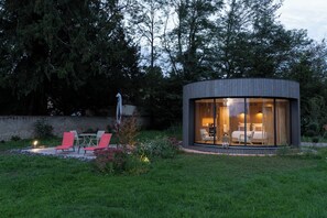 A modern, circular glass structure with a bed and seating area, surrounded by a garden and trees. - Clos Marie (Iguerande)