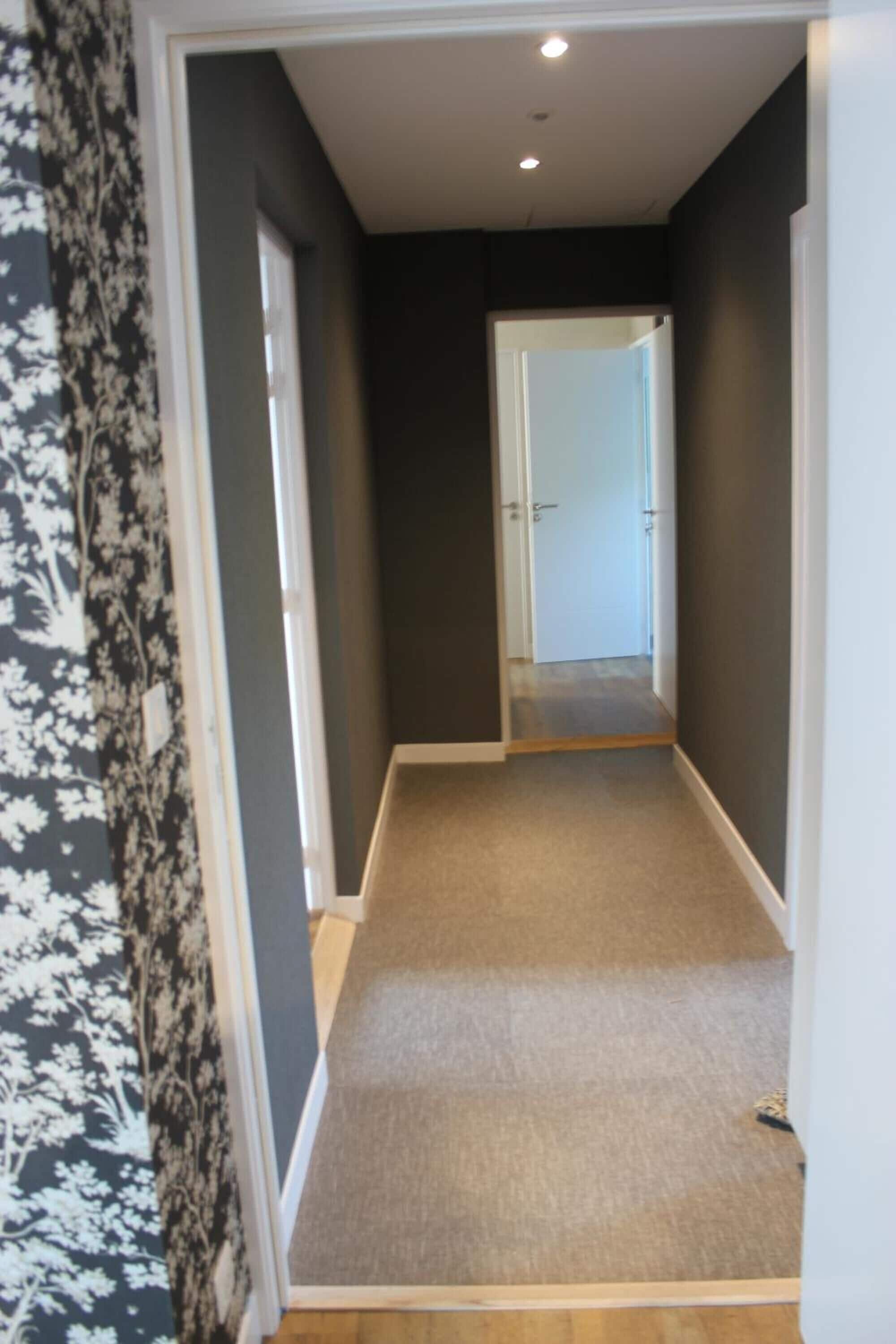 Un estrecho pasillo con suelo alfombrado, una puerta al final y papel tapiz estampado en una pared.