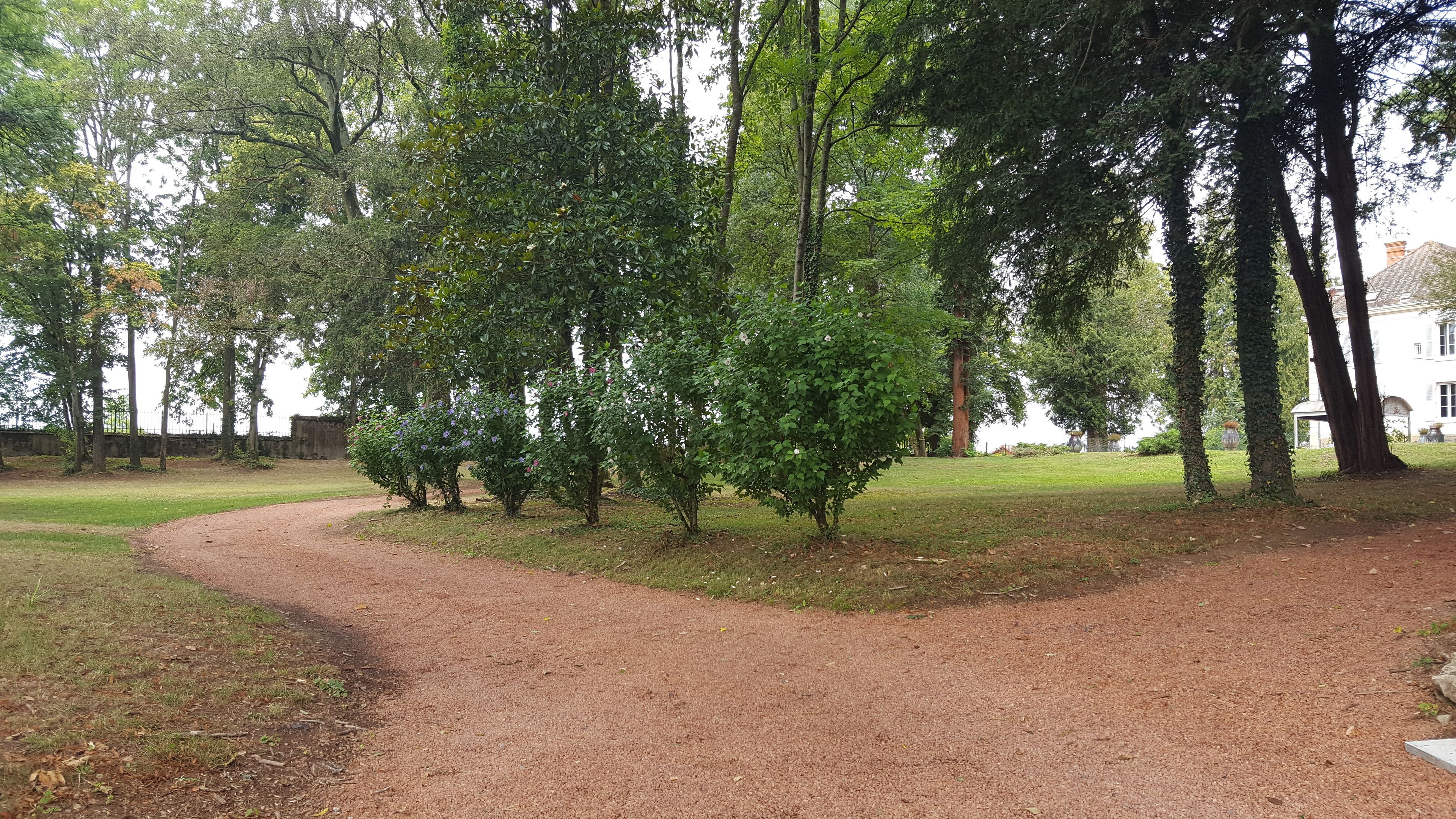 Un sentier de gravier serpente à travers un parc, avec des arbres et un bâtiment blanc en arrière-plan.