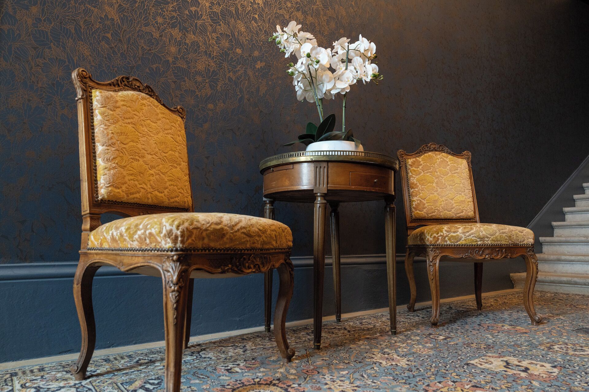 Deux fauteuils richement décorés, dotés de hauts dossiers et de sièges rembourrés, une petite table ronde ornée d’un vase de fleurs blanches et un tapis à motifs.