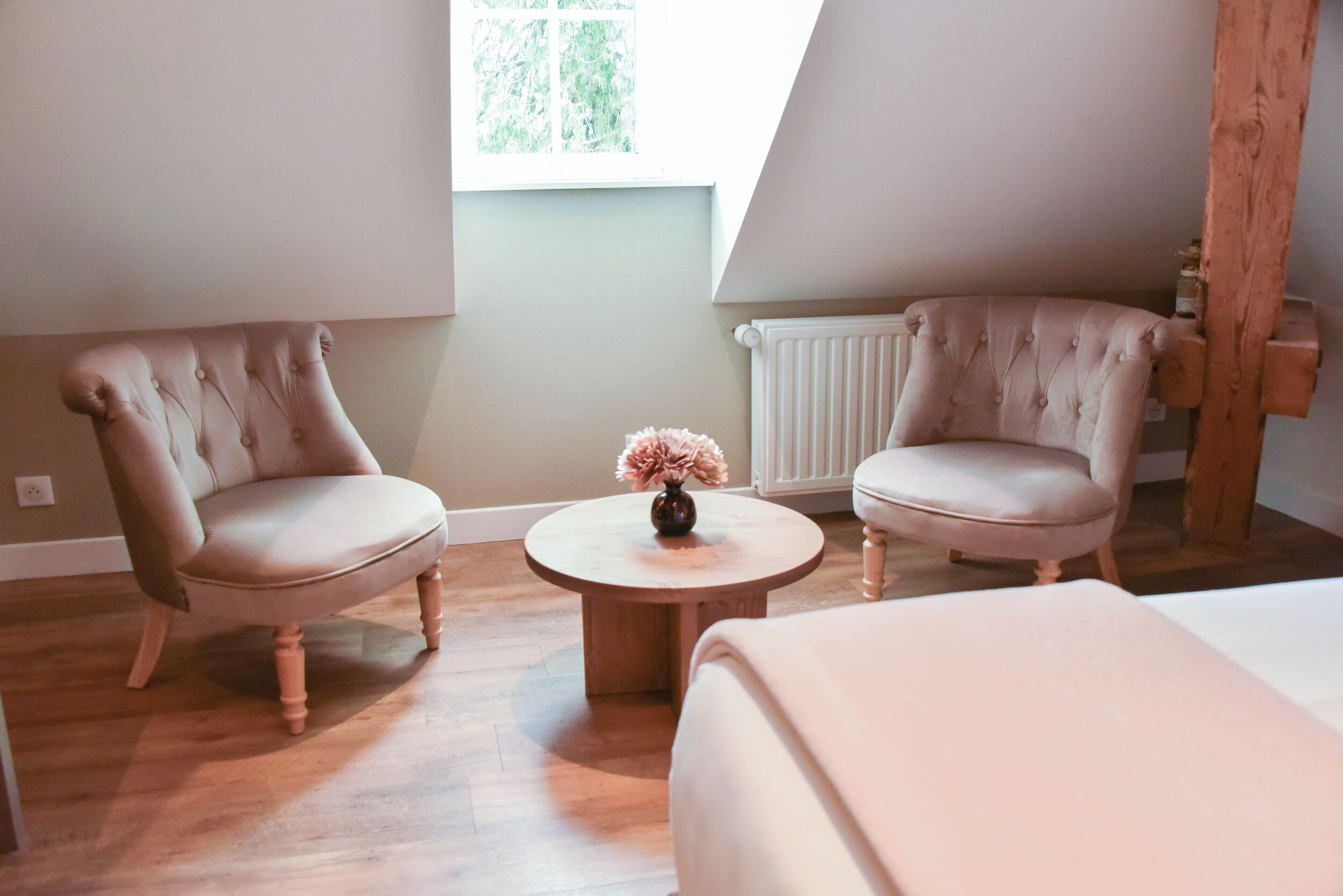 A room with two upholstered chairs, a small round table with a vase, and a bed.
