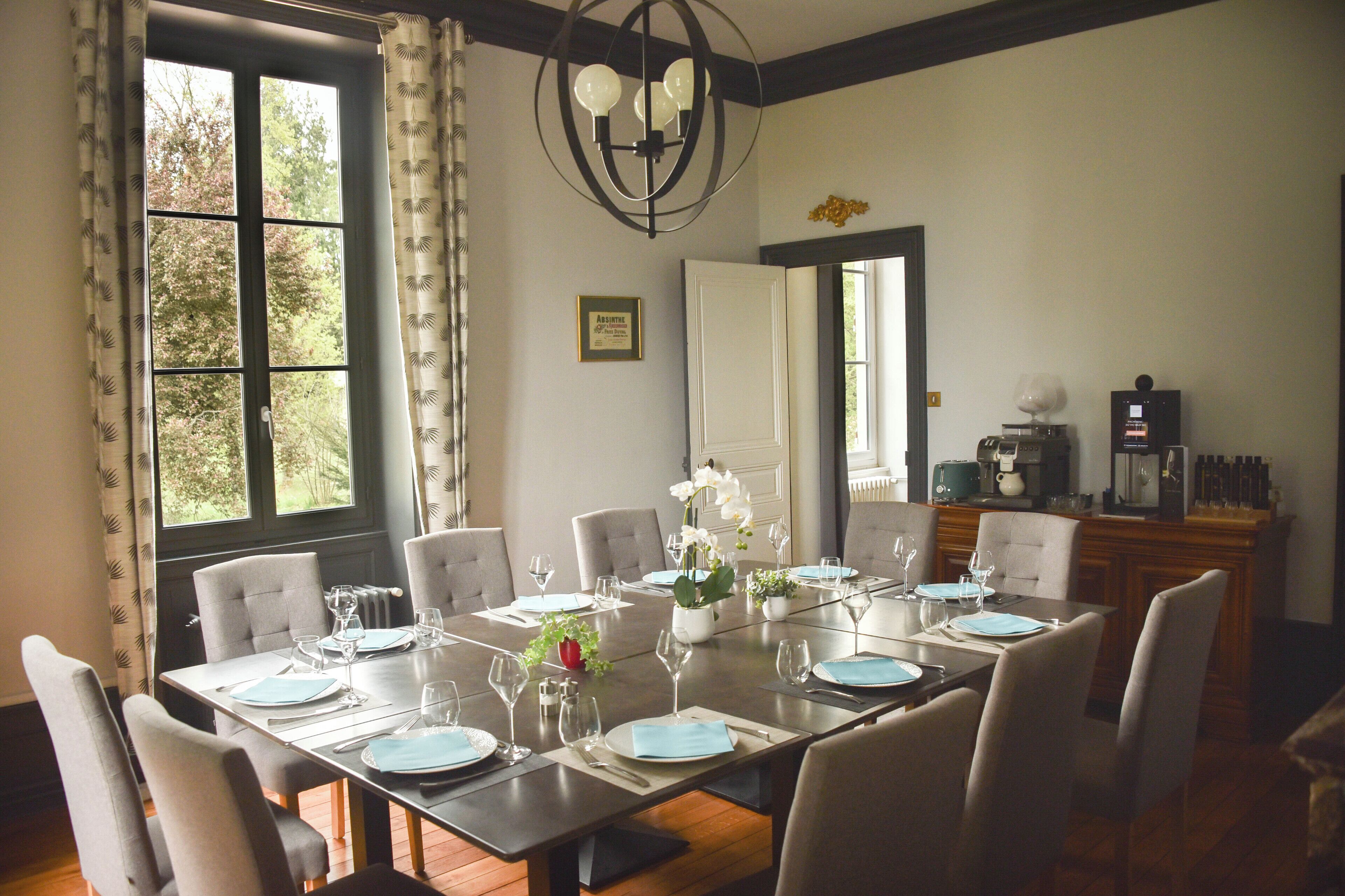 A dining room with a large table set for six, surrounded by chairs. There is a chandelier above the table, and a window with curtains on the left. A cabinet with kitchen appliances is visible in the background.