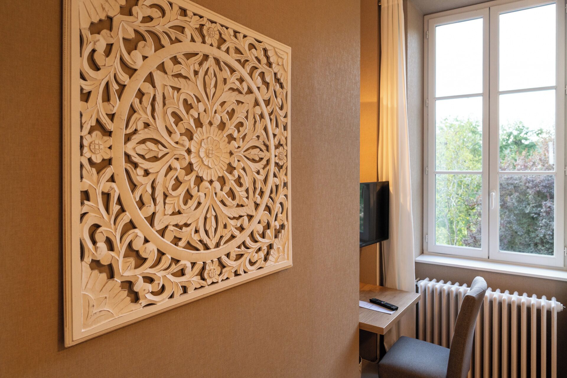 A room with a large, ornate wooden panel on the wall, a window with a view of greenery, a radiator, and a small table with a remote control.
