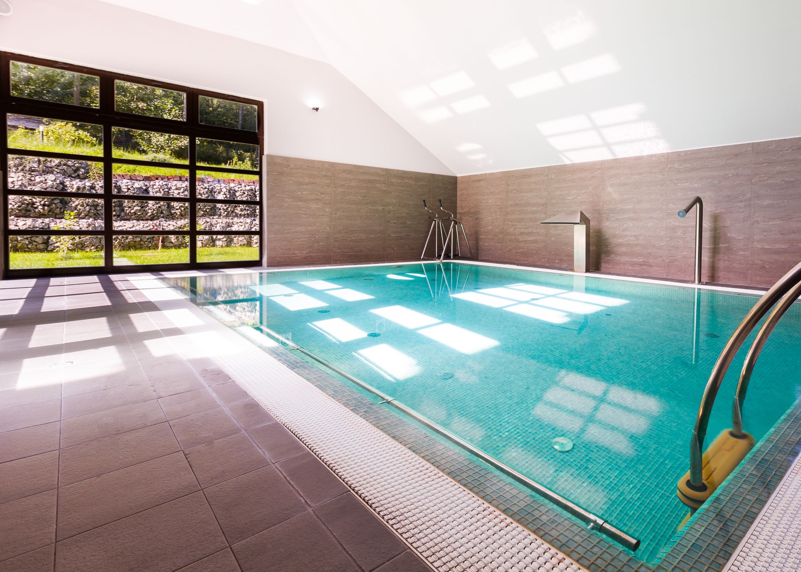 An indoor swimming pool with clear glass walls, a tiled floor, and a view of a garden through large windows.