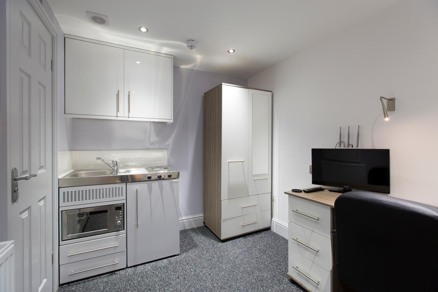 A compact kitchen with a stove, sink, and cabinets, a wooden wardrobe, a television, and a small seating area.