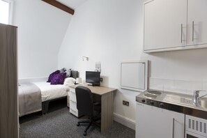 Una habitación pequeña con cama, escritorio, computadora y kitchenette.