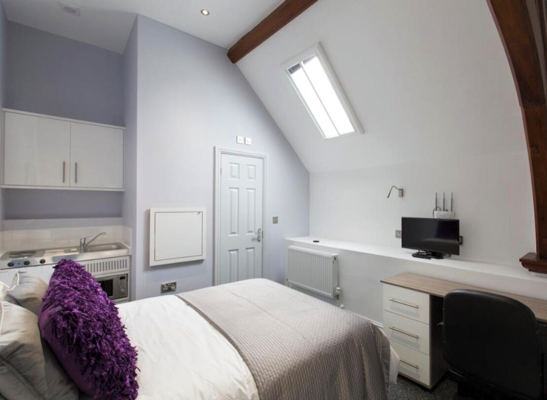 A bedroom with a bed, a desk, a television, a window, and a radiator.