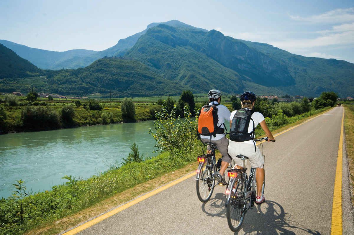 Twee fietsers op een verharde weg, met een rivier en bergen op de achtergrond.