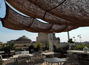 An outdoor seating area with tables and chairs, a woven canopy overhead, and a view of a historic building with a dome in the background. - Lu Leccio (Lecce)