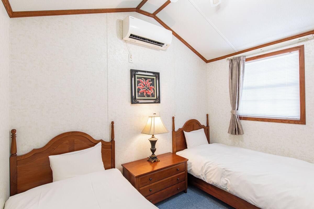 Habitación de hotel con dos camas, una mesita de noche con lámpara, un equipo de aire acondicionado, un cuadro en la pared y una ventana con cortinas.