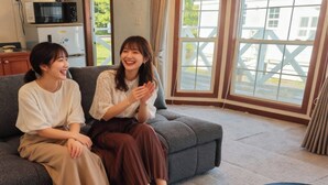Two people sitting on a grey sofa in a room with a large window, a kitchen area with appliances, and a view of greenery outside. - Cottage Star House Nakijin (Nakijin)