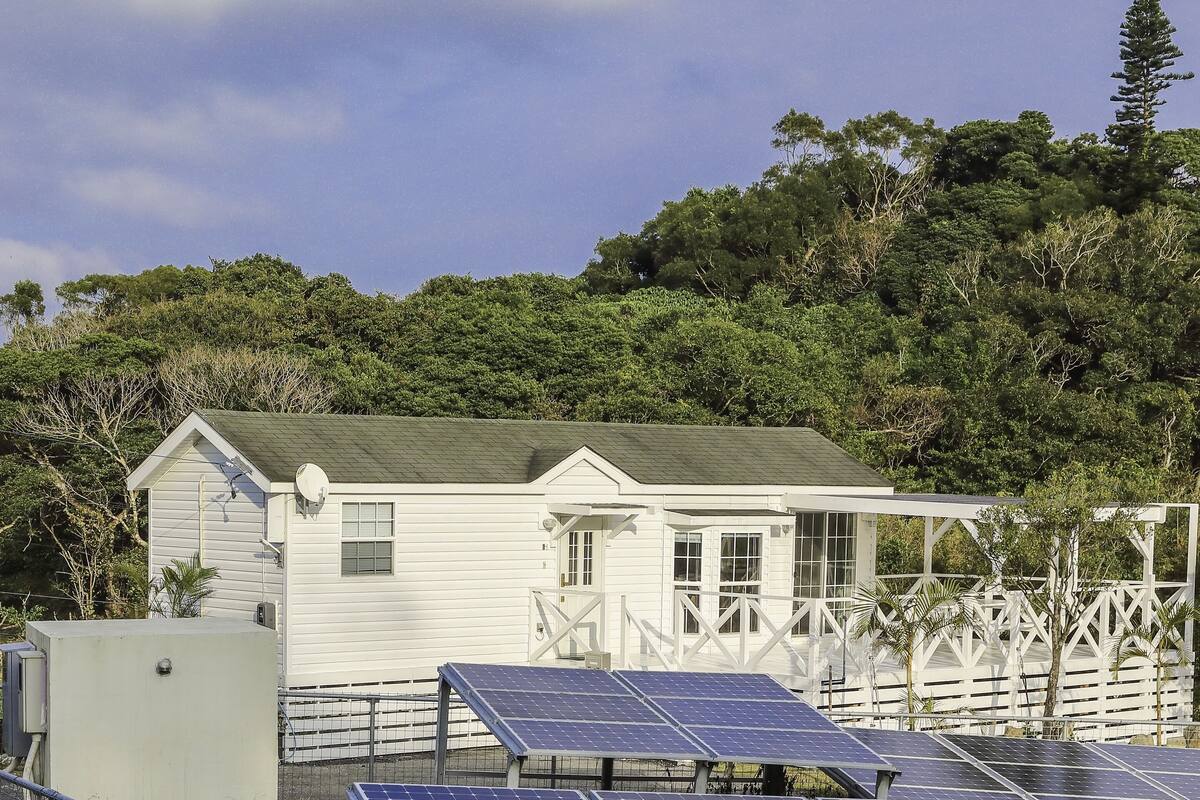 Una casa blanca con techo verde, rodeada de paneles solares y una valla.