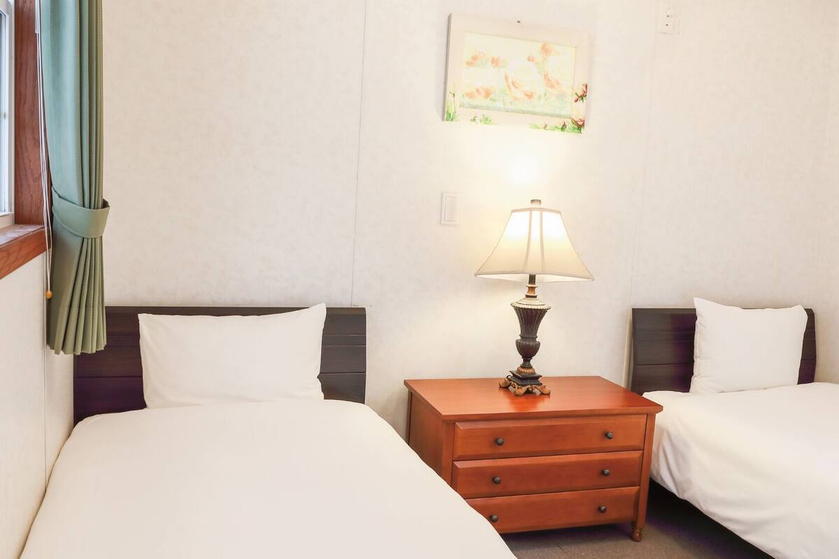 Habitación de hotel con dos camas, una mesita de noche con lámpara, una ventana con cortinas y un cuadro en la pared.