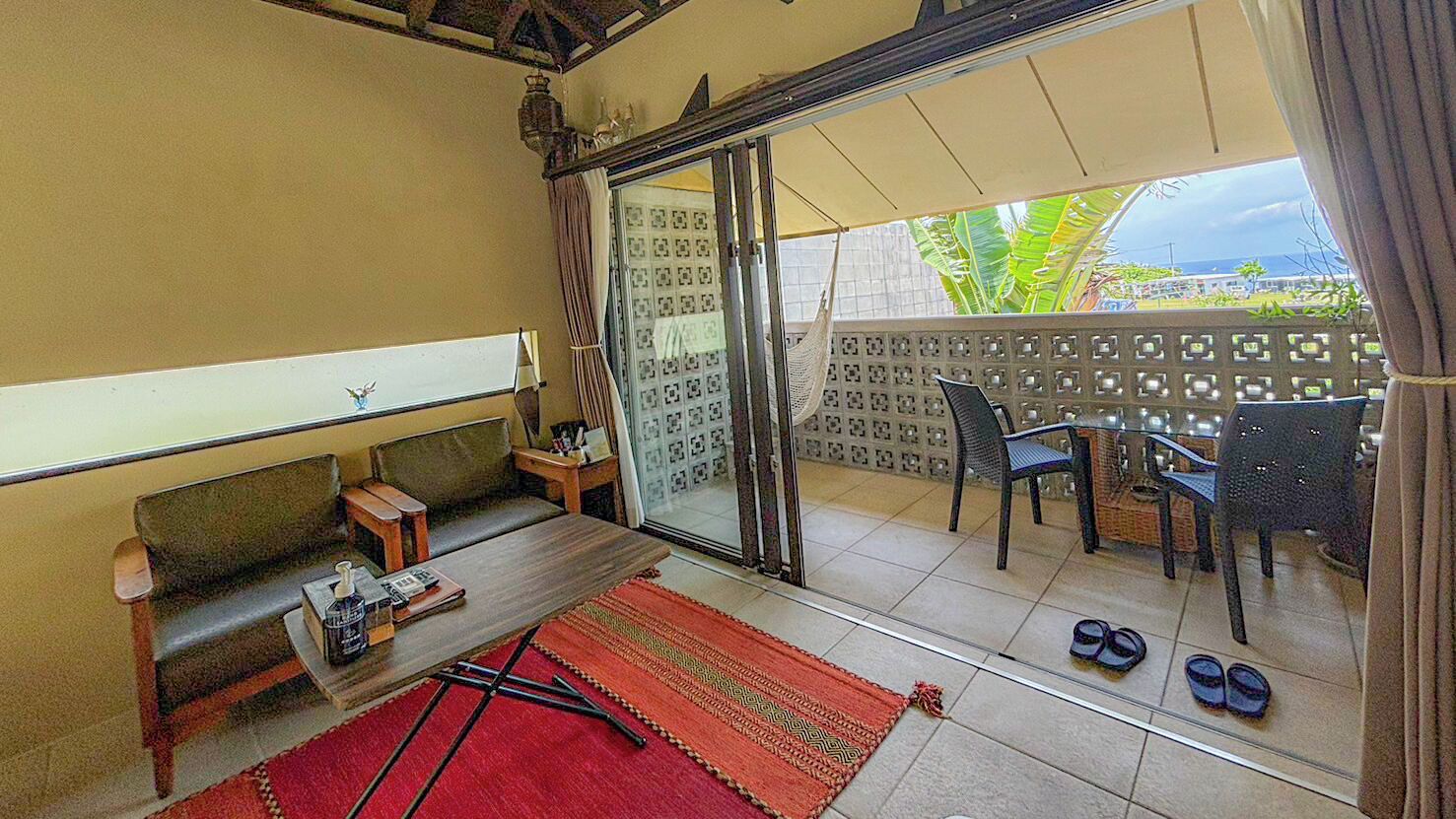 A room with a sofa, a small table with a bottle and a book, a red rug, and a balcony with chairs and a table.