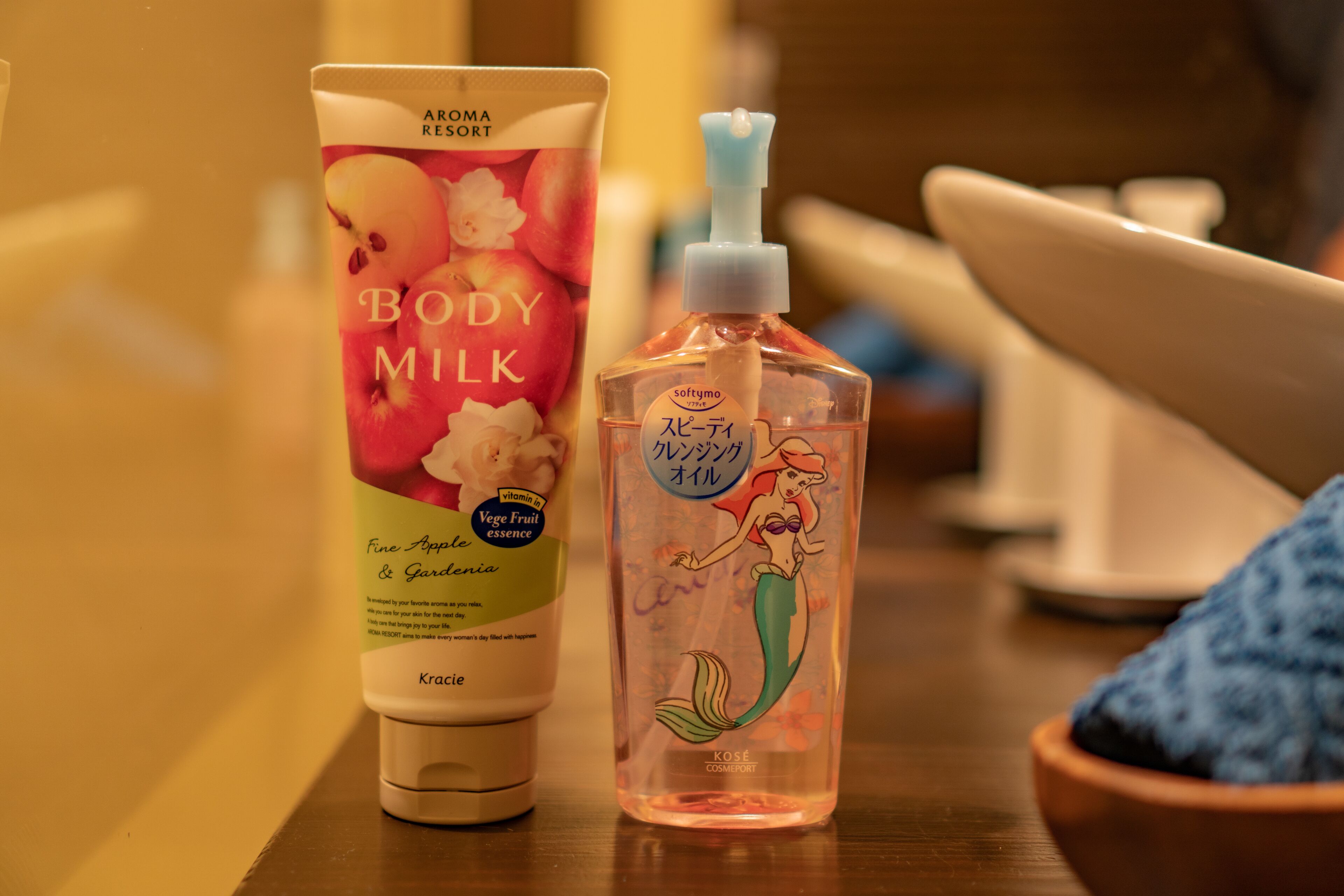 A tube of body milk and a bottle of mermaid-themed body oil on a wooden table.