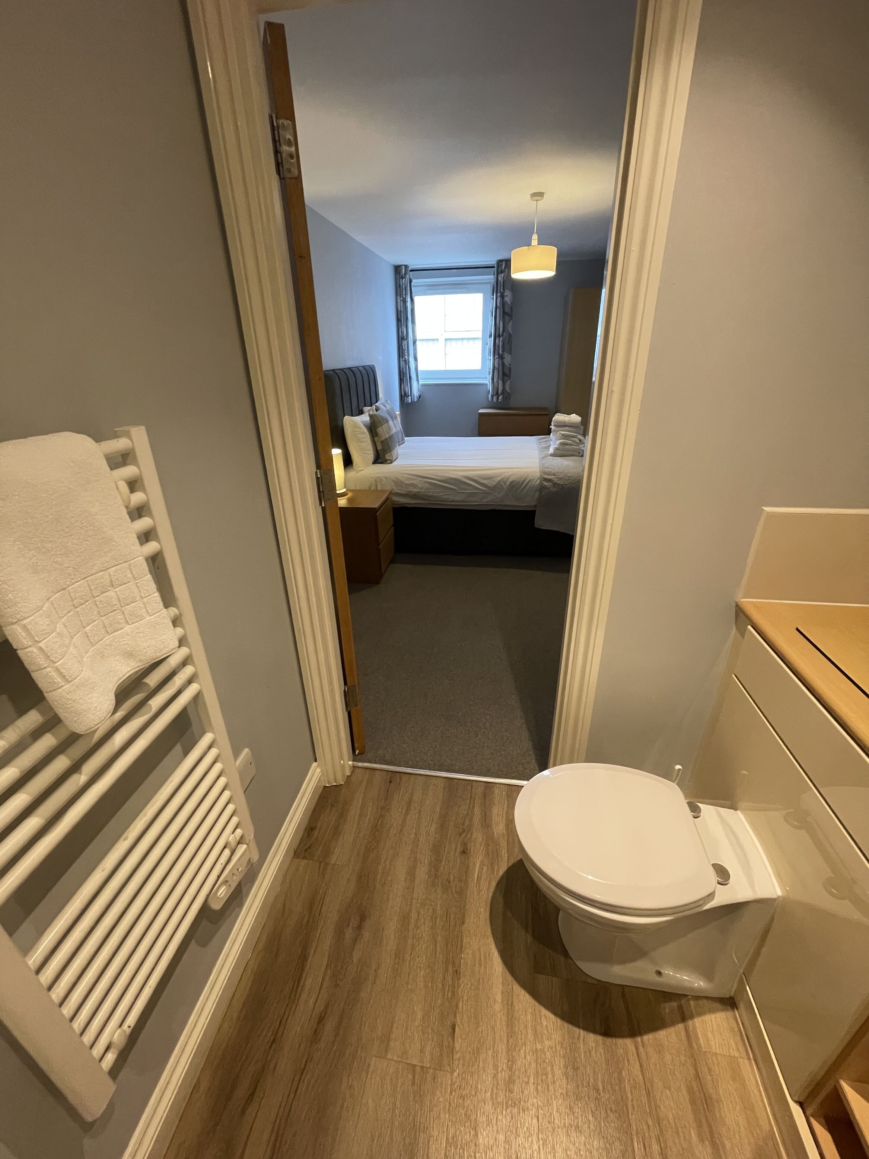 Een kleine slaapkamer met een enkel bed, een nachtkastje, een klein raam en een badkamer met toilet en wastafel.