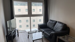 Un salon moderne avec une table basse en verre, un canapé en cuir noir, un téléviseur posé sur un meuble noir et une grande fenêtre donnant sur des immeubles.