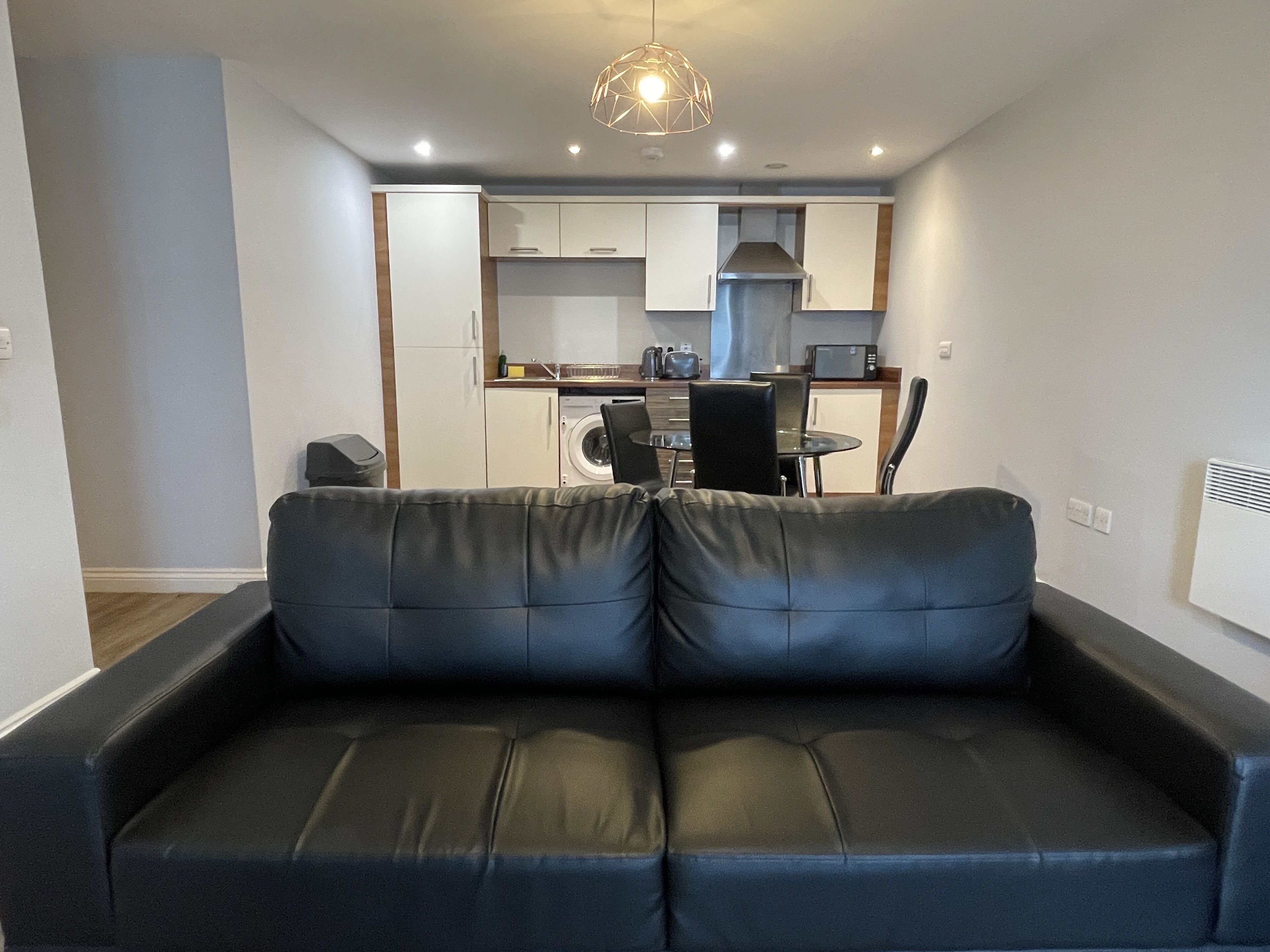 A modern living room with a black leather sofa, a dining area with a table and chairs, and a kitchen in the background.